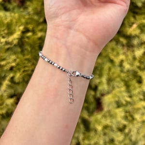 May include: A beaded bracelet with small, round, silver and white beads. The bracelet is on a wrist, and the clasp is visible. The background is a blurred green.