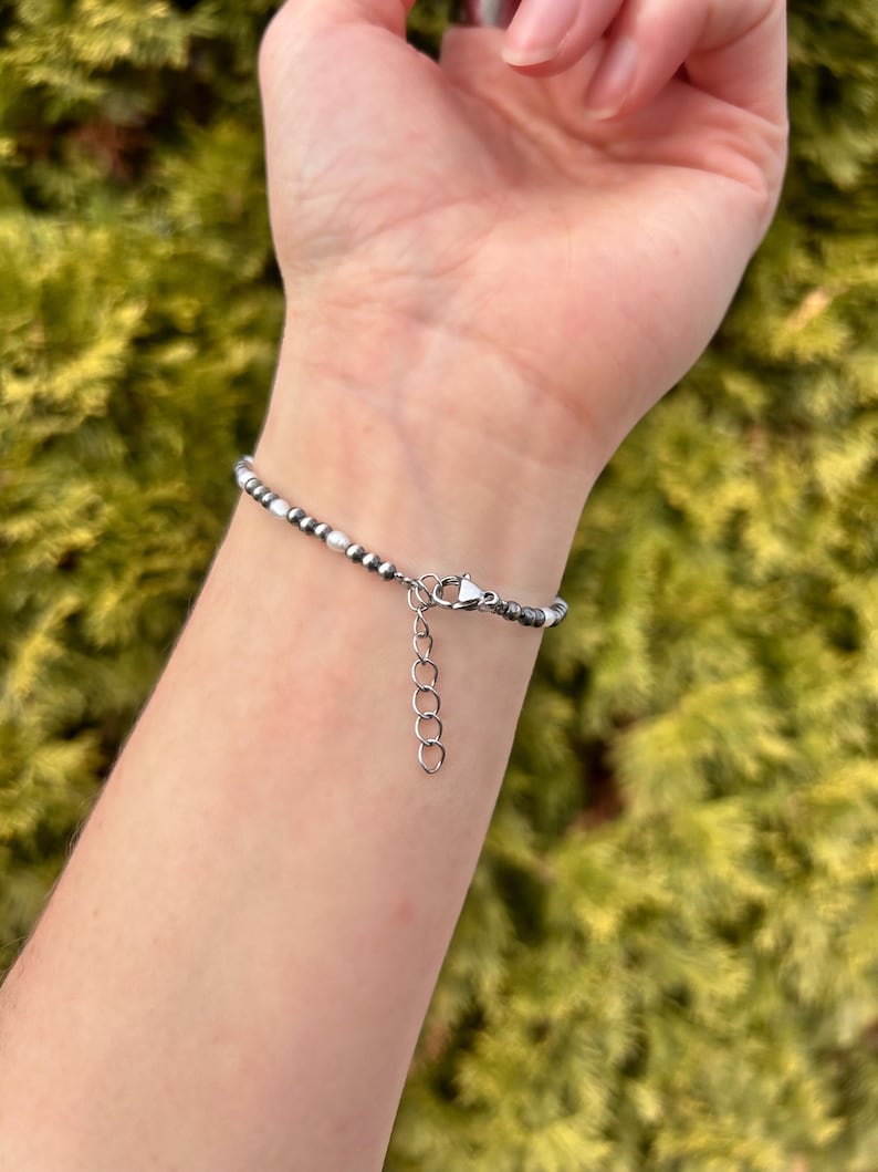 May include: A beaded bracelet with small, round, silver and white beads. The bracelet is on a wrist, and the clasp is visible. The background is a blurred green.