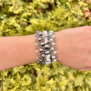 May include: A stack of beaded bracelets in silver, gray, and white. The bracelets feature various bead sizes and textures, including small silver beads, larger gray beads, and white pearls. The bracelets are worn on a wrist.