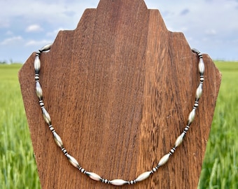 May include: A silver necklace with oval-shaped beads on a wooden display stand against a green grassy field and blue sky background.