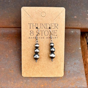May include: A pair of silver earrings with three graduated round beads. The earrings are displayed on a brown card with the text "THUNDER & STONE HANDMADE JEWELRY". The background is a rusty metal surface.