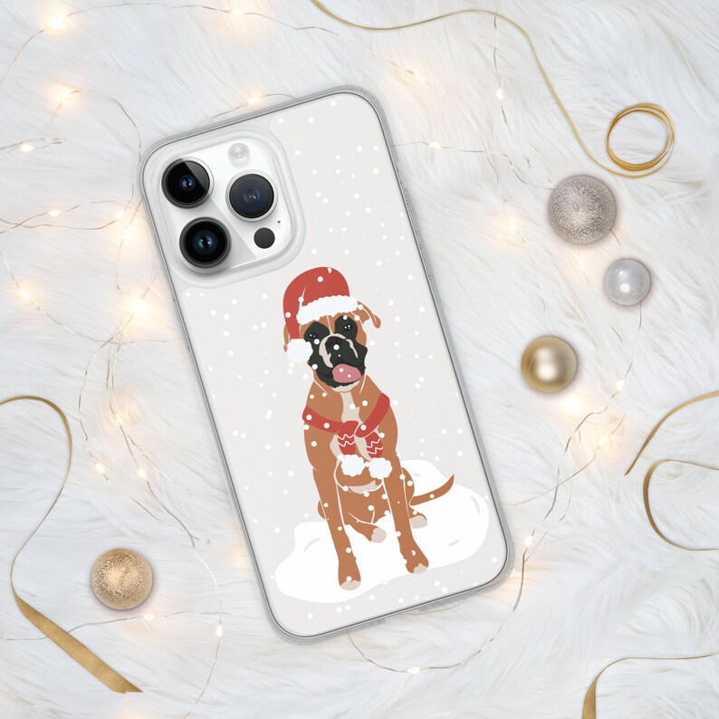 Puede incluir: Una funda transparente para tel&eacute;fono con fondo blanco y un boxer marr&oacute;n y blanco que lleva un gorro de Pap&aacute; Noel rojo y una bufanda. El perro est&aacute; sentado en la nieve con copos de nieve cayendo a su alrededor.