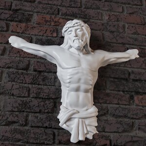 May include: A white sculpture of Jesus Christ on a cross, with arms outstretched. The sculpture has a detailed depiction of the face, hair, and body. The background is a brick wall.
