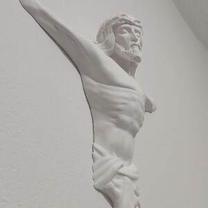 May include: A white plaster sculpture of Jesus Christ on a wall. The sculpture shows Jesus with a crown of thorns, a beard, and outstretched arms. The figure is draped with a cloth around the waist.