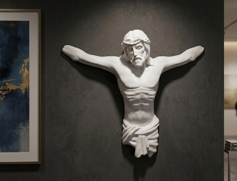 May include: A white sculpture of a figure with outstretched arms, wearing a crown of thorns and a draped cloth. The sculpture is mounted on a dark grey wall. A framed abstract painting is visible on the left.