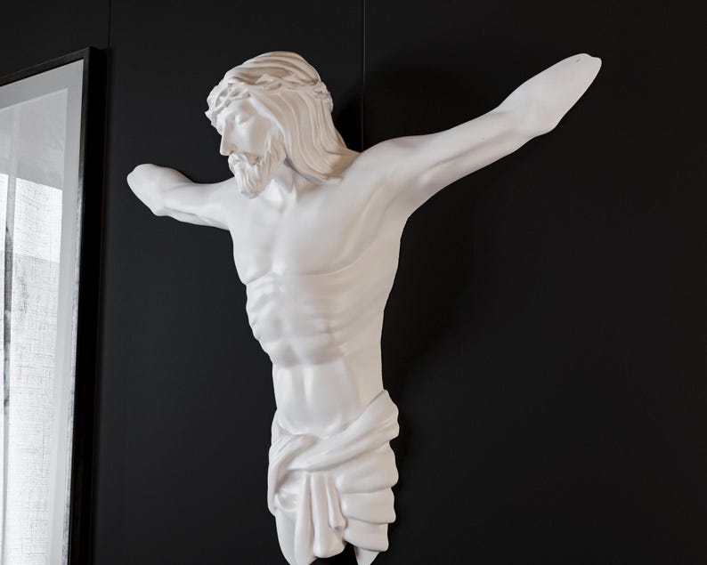 May include: A white sculpture of a man with outstretched arms, a beard, and a crown of thorns, mounted on a black wall. The figure is draped with fabric around the lower body. The sculpture is a religious depiction.
