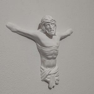 May include: A white plaster sculpture of a man with outstretched arms, wearing a crown of thorns. The figure has a beard and is draped with a cloth around the waist. The sculpture is mounted on a white wall.