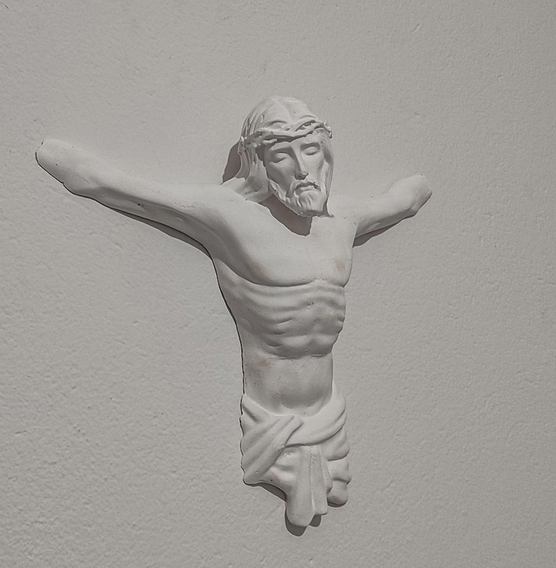 May include: A white plaster sculpture of a man with outstretched arms, wearing a crown of thorns. The figure has a beard and is draped with a cloth around the waist. The sculpture is mounted on a white wall.