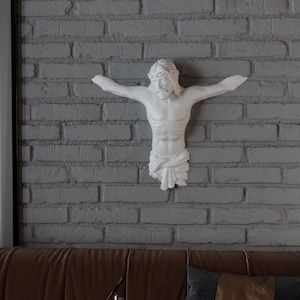 May include: A white sculpture of Jesus Christ on a cross, mounted on a grey brick wall. The sculpture depicts Jesus with outstretched arms and a crown of thorns. The wall is made of grey bricks.