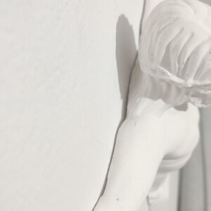 May include: A white plaster sculpture of a figure, possibly an angel or cherub, fixed to a wall. The sculpture features flowing hair and an outstretched arm, creating a shadow on the wall. The overall aesthetic is classical and elegant.