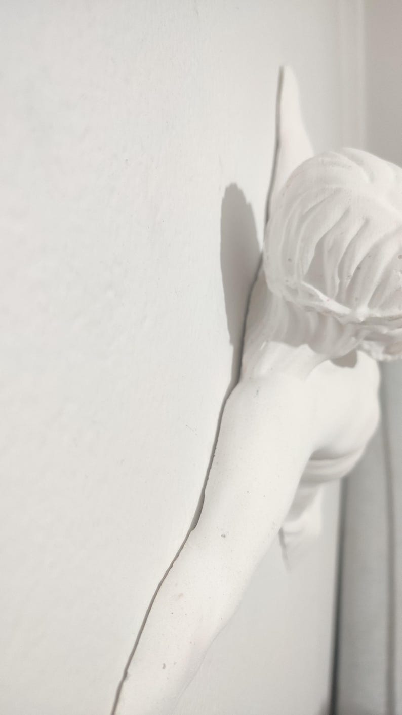May include: A white plaster sculpture of a figure, possibly an angel or cherub, fixed to a wall. The sculpture features flowing hair and an outstretched arm, creating a shadow on the wall. The overall aesthetic is classical and elegant.