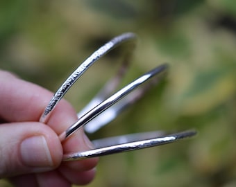 May include: Three silver bangles with a hammered texture. The bangles are stacked on top of each other.