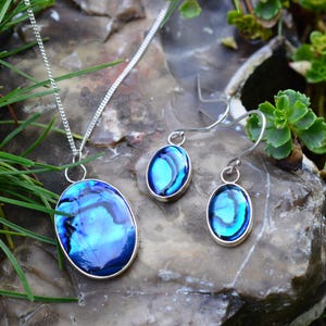 May include: A jewellery set comprising a pendant necklace and matching earrings. The oval pendants and earrings have a vibrant blue and teal iridescent design, set in silver-coloured metal. The necklace chain is also silver. The set is displayed on a stone surface.