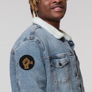 Peut inclure: Veste en jean bleu clair avec col en sherpa blanc. Un écusson circulaire noir avec un poing doré et un motif floral est sur la manche gauche. La veste a deux poches poitrine et une fermeture boutonnée.