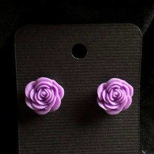 Könnte beinhalten: Ein Paar violette Rosen-Ohrstecker. Die Ohrringe sind aus einem weichen, biegsamen Material gefertigt und haben ein realistisches Rosen-Design.