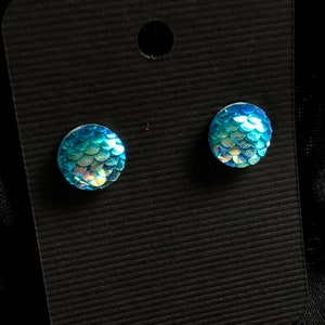 Puede incluir: Un par de pendientes de botón azul iridiscente con un diseño de escamas de sirena.