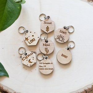 Puede incluir: Siete chapas para perros de madera con nombres y diseños grabados. Las chapas están sobre una superficie de madera. Los nombres en las chapas son Luna, Loki, Scout, Dawn, Navi, Bandit y 123 Springdale St. Zion, UT (435)-555-8095.