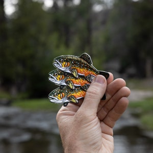 May include: Three embroidered patches of trout fish in shades of green, orange, and yellow. The patches are held in a hand against a blurred background of trees and a stream.