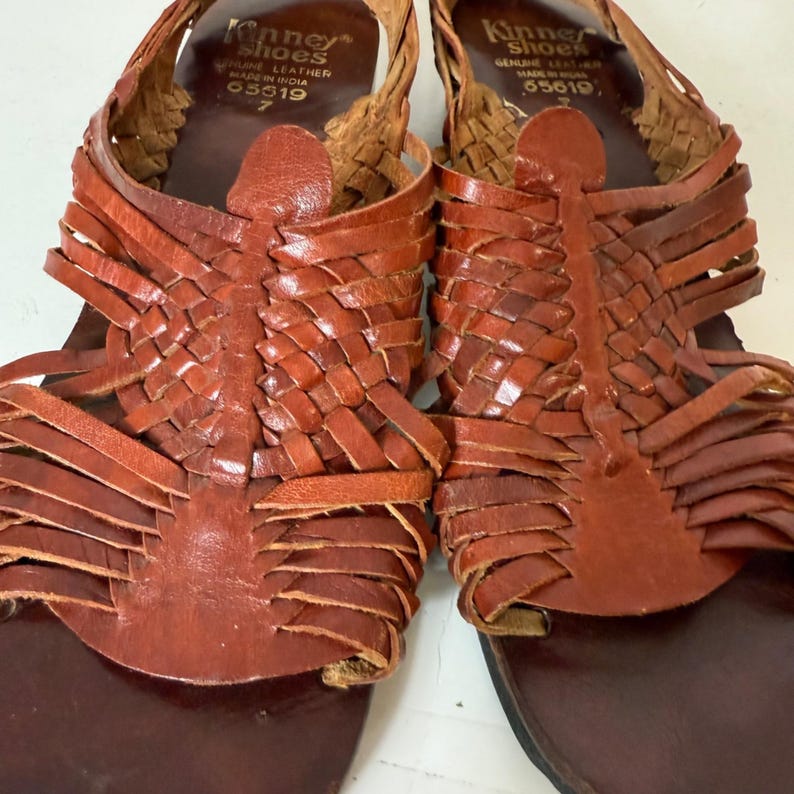 May include: Pair of brown leather sandals with woven straps and a dark brown sole. The sandals have a unique design with intricate weaving across the top and around the ankle. The brand name "Kinney Shoes" is visible on the insole.