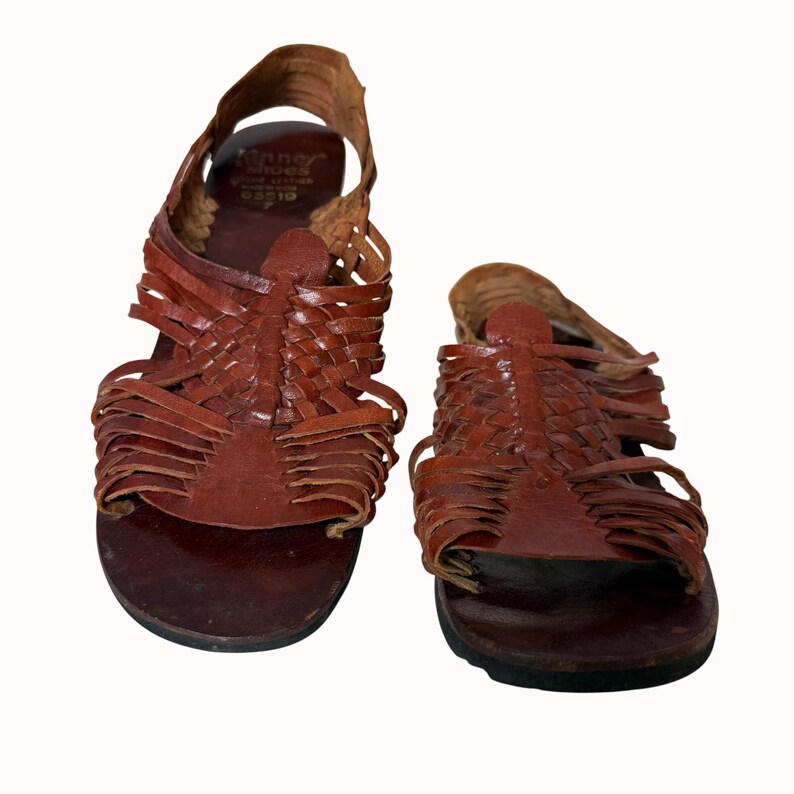 May include: A pair of brown leather sandals with woven straps and a slingback design. The sandals have a dark brown sole and a textured appearance. The brand name "Kenner Shoes" is visible on the left sandal.