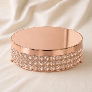 May include: A round cake stand with a reflective rose gold metallic top surface. The side of the stand features a decorative lattice design embedded with numerous clear, circular crystals, supported by small, rounded metallic feet.