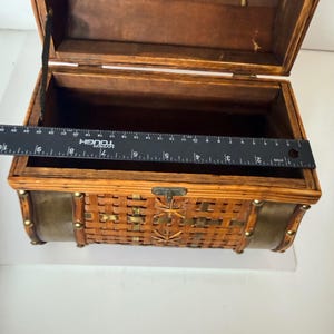 May include: Open wooden chest with a woven front and brass accents. The chest has a hinged lid and a metal clasp. A ruler is placed inside the chest, measuring approximately 10 inches across.