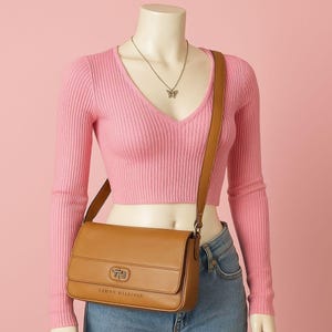 May include: A tan Tommy Hilfiger crossbody bag with a rectangular shape and a flap closure. The bag has a long, adjustable strap and a silver-tone clasp. The brand name is visible on the front of the bag.