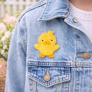 Puede incluir: Un parche de pato bordado amarillo está fijado a una chaqueta vaquera. El pato tiene un aspecto texturizado con ojos negros y patas naranjas. La chaqueta es azul claro con botones color latón.