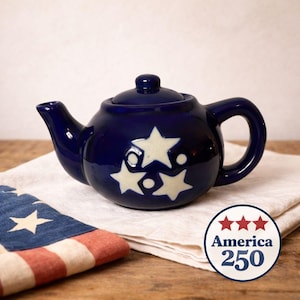 Puede incluir: Una tetera de cerámica azul marino con tapa y asa, decorada con motivos de estrellas color crema. La tetera está colocada sobre un paño blanco, con una pequeña bandera americana y un logotipo de "America 250" en el fondo.