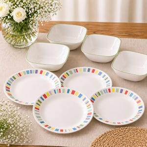 May include: A collection of white ceramic plates and bowls. The plates feature a colourful striped border in red, yellow, blue, and green. The bowls are square with a green trim. The set is displayed on a neutral tablecloth.