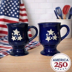 May include: Two navy blue ceramic mugs with white star designs. The mugs have a pedestal base and curved handles. An American flag and a red, white, and blue patterned cloth are in the background. A circular emblem with the text "AMERICA 250" is visible.