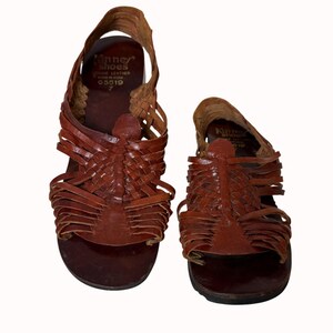 May include: Pair of brown leather sandals with woven straps. The sandals have a flat sole and a strap that goes around the ankle. The brand name "Kinney Shoes" is visible on the insole.