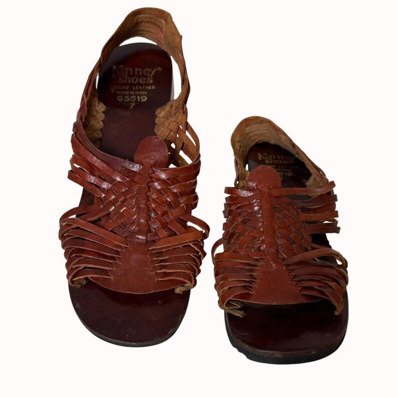 May include: Pair of brown leather sandals with woven straps. The sandals have a flat sole and a strap that goes around the ankle. The brand name "Kinney Shoes" is visible on the insole.