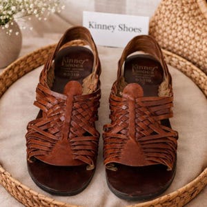 May include: A pair of brown leather sandals with woven straps. The sandals have a slingback design and a dark brown sole. The brand name "Kinney Shoes" is visible on a tag in the background. The sandals are displayed on a woven tray.