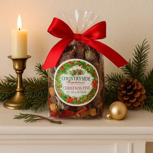 以下が含まれることがあります： 赤いリボンで結ばれた、ポプリが入った透明なセロハン袋。「Countryside Gingerbreads Christmas Pine」と書かれた丸いラベル。真鍮の燭台のろうそく、松の枝、松ぼっくり、金のオーナメントが、クリスマスのシーンを完成させています。