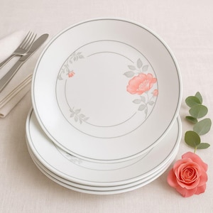 May include: Four white ceramic plates with a grey border and floral design. Each plate has a pink flower and grey leaves. A fork, knife, and napkin are in the background, with a pink rose and greenery to the side.