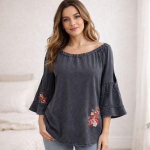 May include: Dark gray off-the-shoulder top with three-quarter sleeves. The sleeves have a flared design with embroidered floral accents in red, pink, and white. The top has a relaxed fit and a slightly faded appearance.