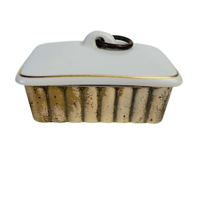 以下が含まれることがあります： 金色の縁取りと茶色の木製ハンドルが付いた白い陶器製のバター入れ。バター入れは、質感のある金色の底面をしています。