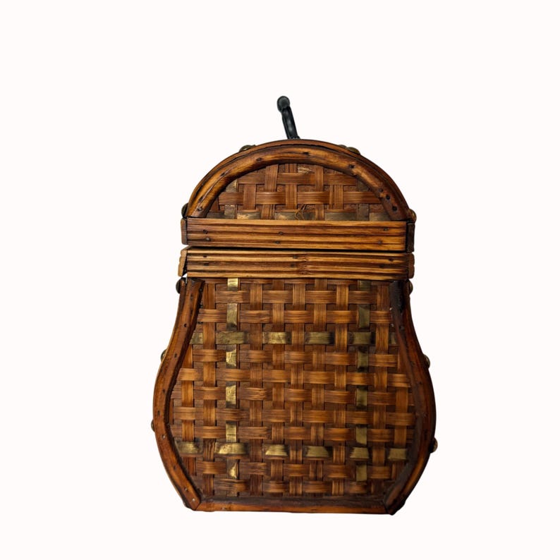 May include: A woven bamboo box with a curved top and a black handle. The box is a warm brown color with brass accents and a woven pattern. The box has a rectangular shape with rounded edges.
