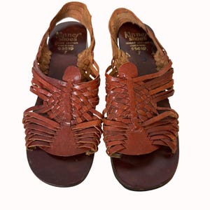 May include: A pair of brown leather sandals with woven straps. The sandals have a rounded toe and a strap that goes around the ankle. The brand name "Kinney Shoes" is visible on the inner side of the sandal. Size 7.