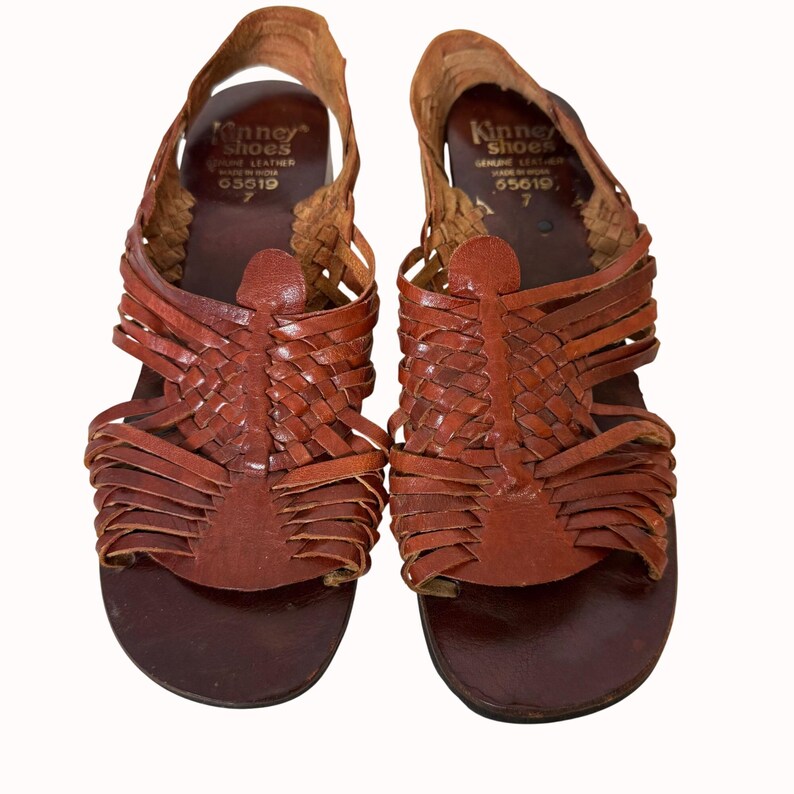 May include: A pair of brown leather sandals with woven straps. The sandals have a rounded toe and a strap that goes around the ankle. The brand name "Kinney Shoes" is visible on the inner side of the sandal. Size 7.