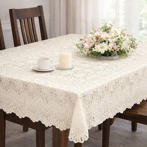 May include: A rectangular cream-colored crochet tablecloth featuring an intricate floral and scalloped lace pattern. The tablecloth is draped over a table, topped with a white ceramic mug, a cylindrical pillar candle, and a floral arrangement.
