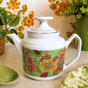 May include: A white ceramic teapot featuring a prominent green band around the centre decorated with a repeating pattern of orange and brown stylised flowers and leaves. The teapot includes a matching lid with a knob handle and gold trim.