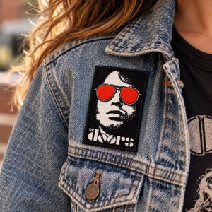 Könnte beinhalten: Ein rechteckiger gestickter Aufnäher mit einem schwarz-weißen Porträt einer Person, die eine leuchtend rote Fliegerbrille trägt. Der Text "THE DOORS" ist in Weiß am unteren Rand des Aufnähers aufgedruckt, der an einer blauen Jeansjacke befestigt ist.
