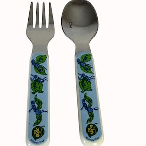 May include: A stainless steel fork and spoon set for children. The handles are light blue with a repeating pattern of green leaf and insect illustrations. The words "bugs life" and the Disney/Pixar logo are printed on the handles.