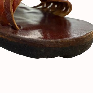 May include: Close-up of a brown leather sandal. The sandal features a dark brown sole and a reddish-brown leather strap. The leather appears worn, suggesting a vintage or handcrafted item. The sandal is designed for comfort and style.