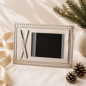 May include: A rectangular silver-toned picture frame featuring a textured beaded border and a central black display area. The left side of the frame is adorned with a raised, crossed ski emblem, while the inner border is lined with clear crystals.