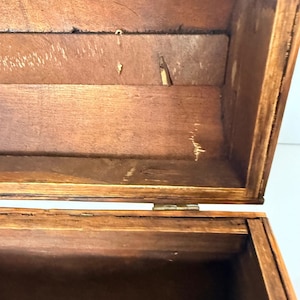 May include: Open wooden box with a hinged lid, revealing the interior. The box is constructed from stained wood, showcasing the natural grain and texture. The interior shows signs of wear and tear, adding to its vintage appeal.