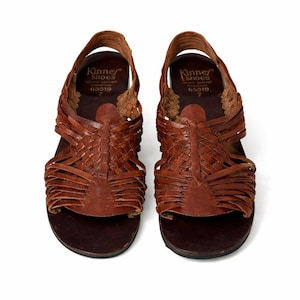 May include: Pair of brown leather sandals with woven straps. The sandals have a slingback design and a dark brown sole. The brand name "Kinney Shoes" is visible on the insole. Size 7.