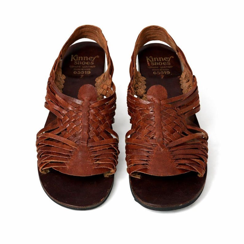 May include: Pair of brown leather sandals with woven straps. The sandals have a slingback design and a dark brown sole. The brand name "Kinney Shoes" is visible on the insole. Size 7.
